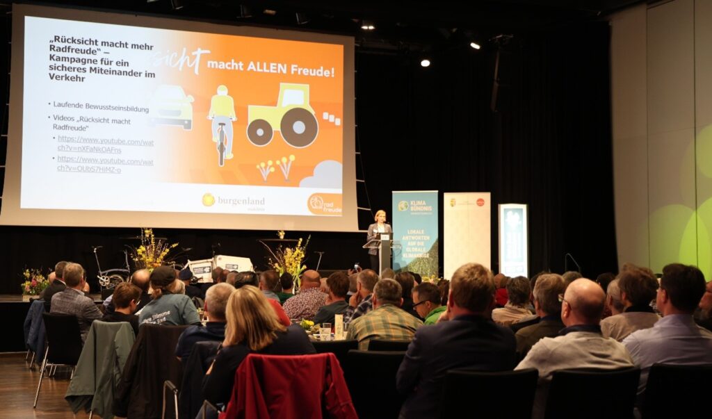 Publikum im Vordergrund, Rednerin auf Bühne mit Präsentationsleinwand